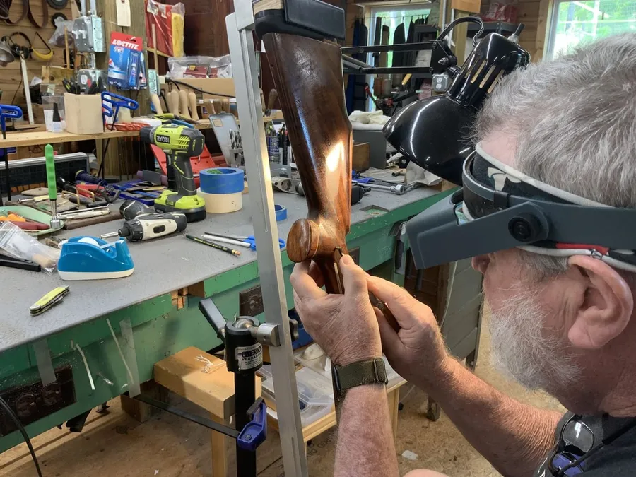 David in profile, wearing a magnifier visor, holding a finished walnut stock up to the window light